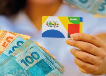 Cartão do Bolsa Família sendo segurado com cédulas de dinheiro visíveis ao fundo, representando o benefício financeiro para famílias de baixa renda
