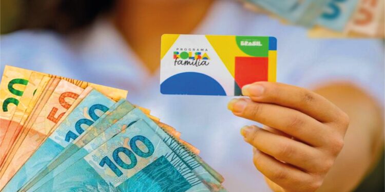 Cartão do Bolsa Família sendo segurado com cédulas de dinheiro visíveis ao fundo, representando o benefício financeiro para famílias de baixa renda