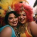 Duas mulheres sorridentes em trajes carnavalescos coloridos, com penas e acessórios dourados, aproveitando a folia