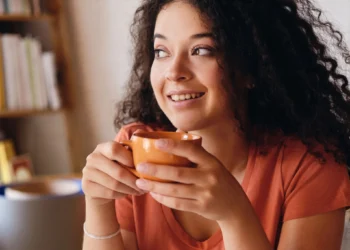 Mulher sorrindo enquanto segura uma xícara de chá, em ambiente caseiro e aconchegante.