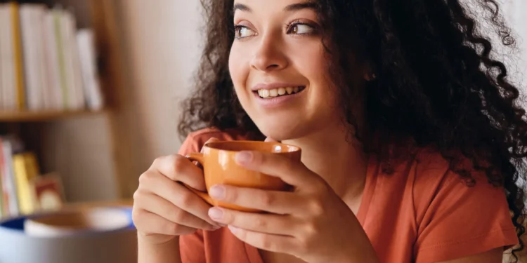 Mulher sorrindo enquanto segura uma xícara de chá, em ambiente caseiro e aconchegante.