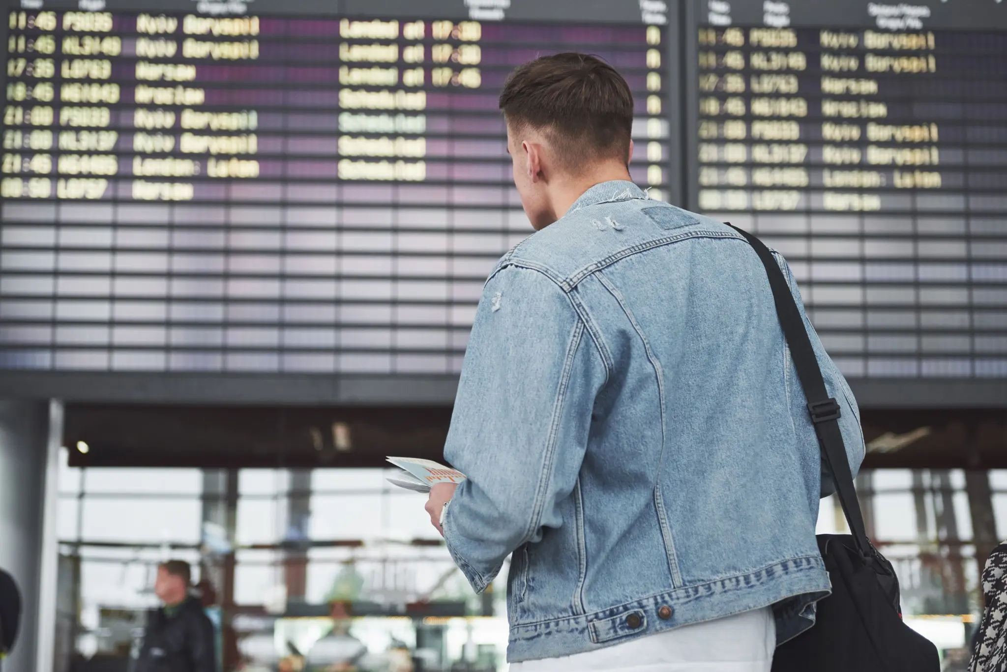 Homem observando o painel de voos no aeroporto, segurando um bilhete de viagem enquanto aguarda o embarque.