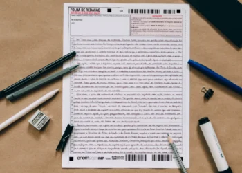 Folha de Redação do Enem, mostrando a escrita manuscrita de um candidato com canetas e lápis ao redor.