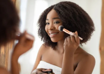 Mulher sorrindo enquanto aplica maquiagem no rosto com um pincel, em um ambiente bem iluminado. Dicas de beleza rápida.