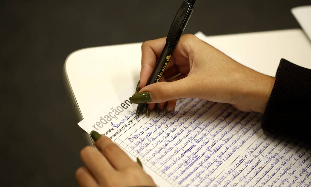 Pessoa escrevendo a redação do ENEM com caneta, com foco na folha de redação manuscrita.
