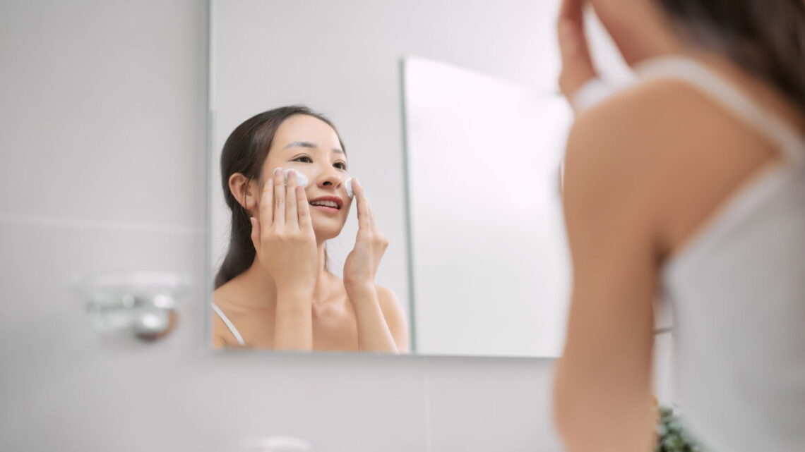 Mulher aplicando um produto de limpeza no rosto enquanto sorri, refletida em um espelho.